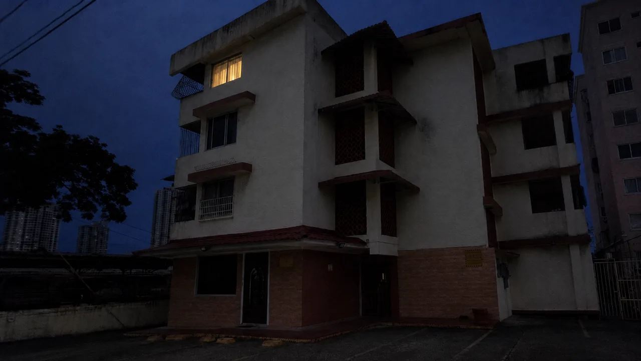 Un edificio de apartamentos de noche, visto desde la calle, con una sola ventana encendida.